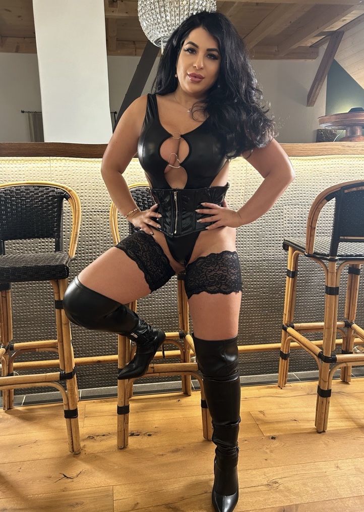 Lady Ayse lehnt an einer Bar in Corsage und hohen Leder Stiefeln dominant und streng